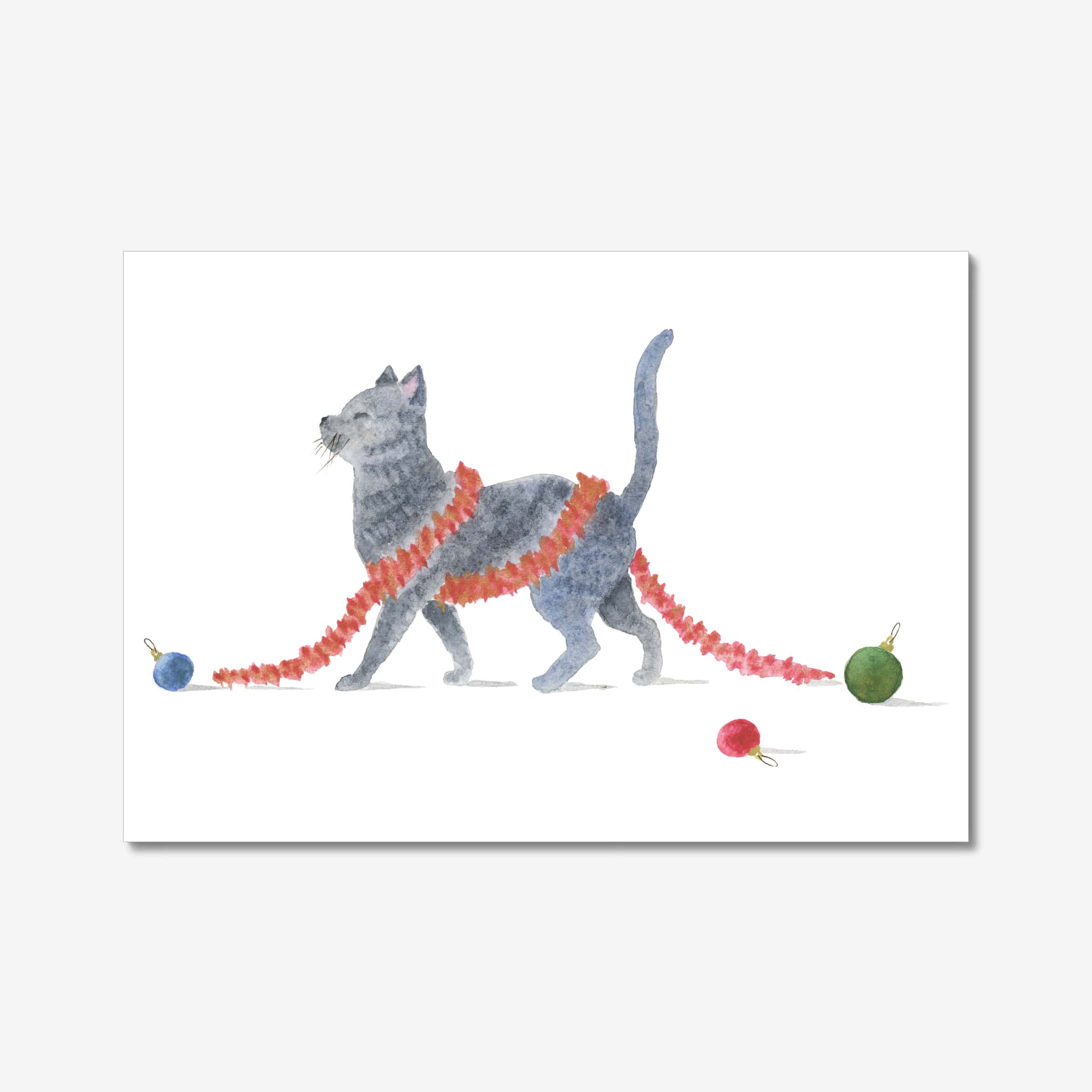 Kissakortti jouluksi – mustanharmaa kissanpentu kulkee kuusenkoristenauha ympärillään ja koristepallot lattialla